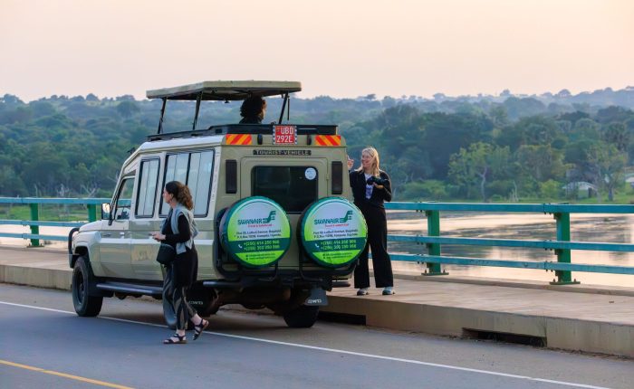 Tourists taking photos outside a Swanair 4x4 pop-top safari Land Cruiser stopped on a scenic bridge over a wide river.