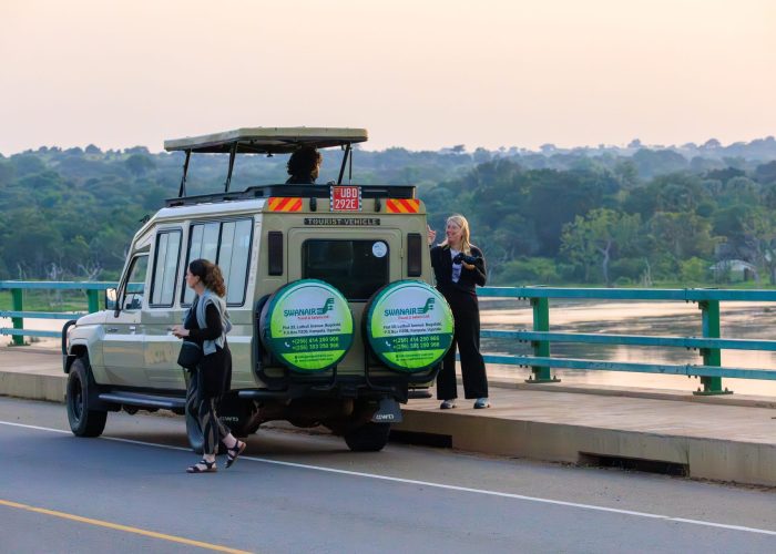 Tourists taking photos outside a Swanair 4x4 pop-top safari Land Cruiser stopped on a scenic bridge over a wide river.