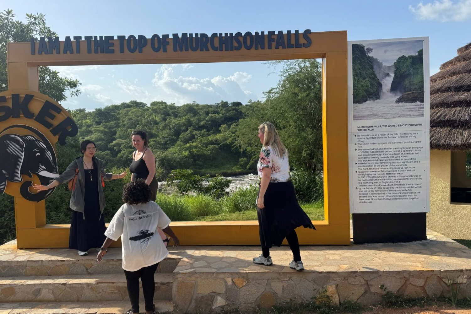 A group of tourists posing and laughing in front of the large yellow 'Top of Murchison Falls' photo frame monument.