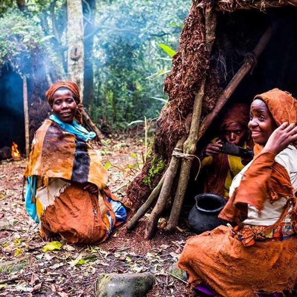 Batwa community members wearing traditional barkcloth garments sitting happily outside thatched forest shelters.