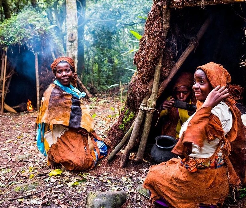 Batwa community members wearing traditional barkcloth garments sitting happily outside thatched forest shelters.