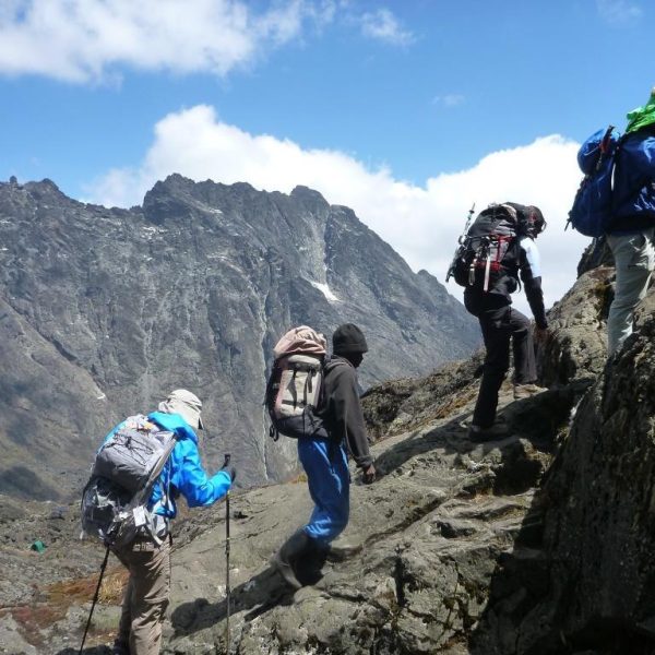 A group of hikers with backpacks and trekking poles scaling a steep, rocky ridge in the Rwenzori Mountains against dramatic peaks.