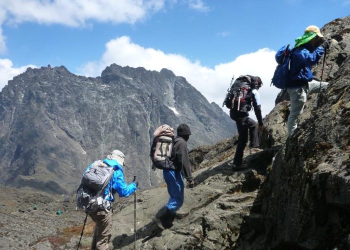 A group of hikers with backpacks and trekking poles scaling a steep, rocky ridge in the Rwenzori Mountains against dramatic peaks.
