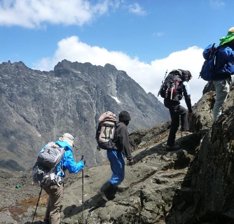 A group of hikers with backpacks and trekking poles scaling a steep, rocky ridge in the Rwenzori Mountains against dramatic peaks.