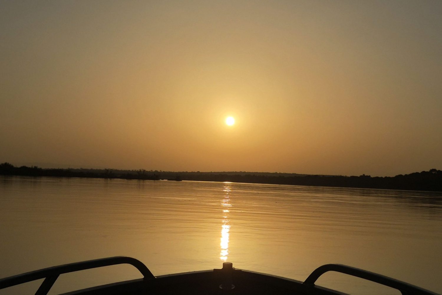A golden sunset reflecting across calm river waters, viewed from the bow of a safari boat cruising along the Nile.